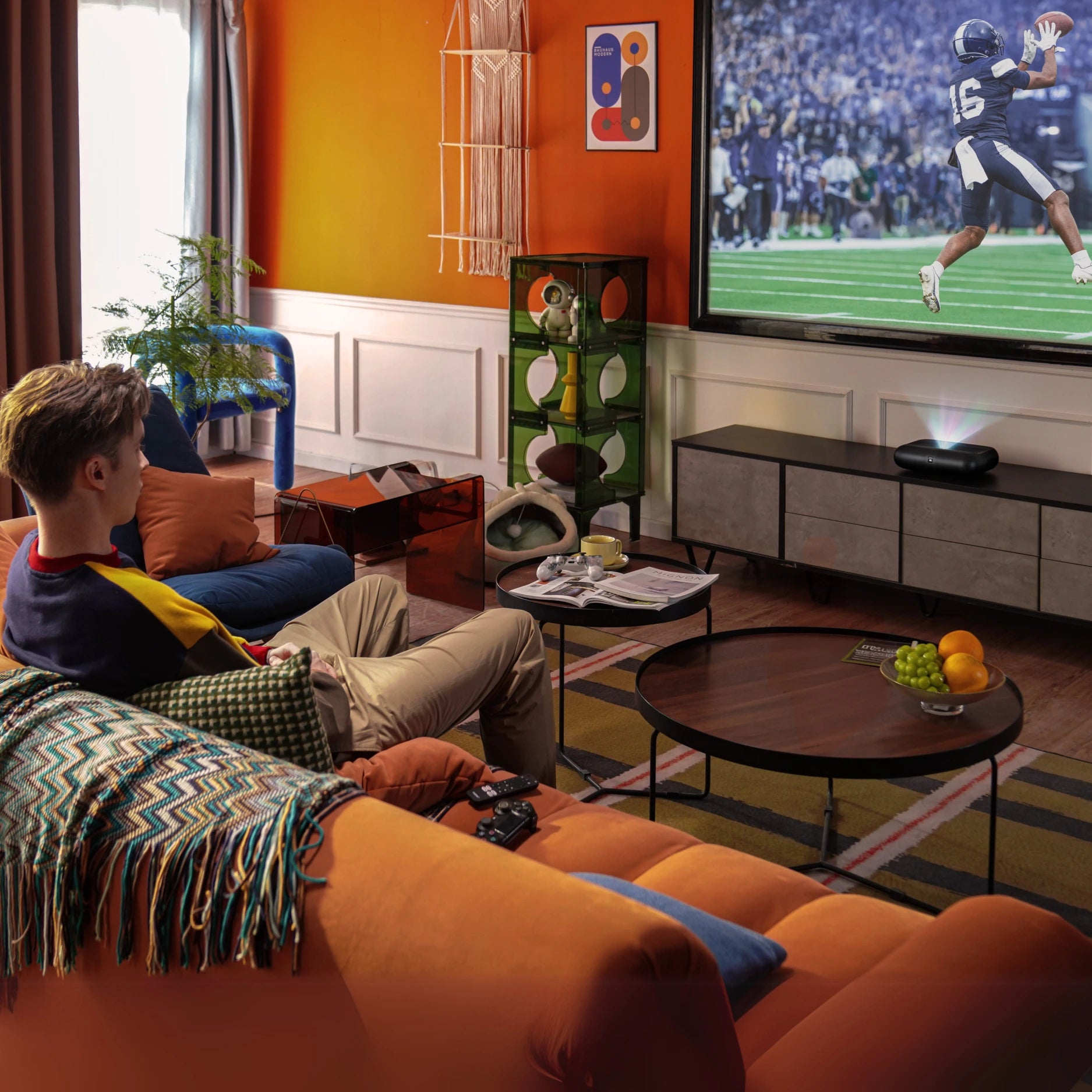 Living room with a person watching on a laser projector displaying a sports game, surrounded by furniture and decor.