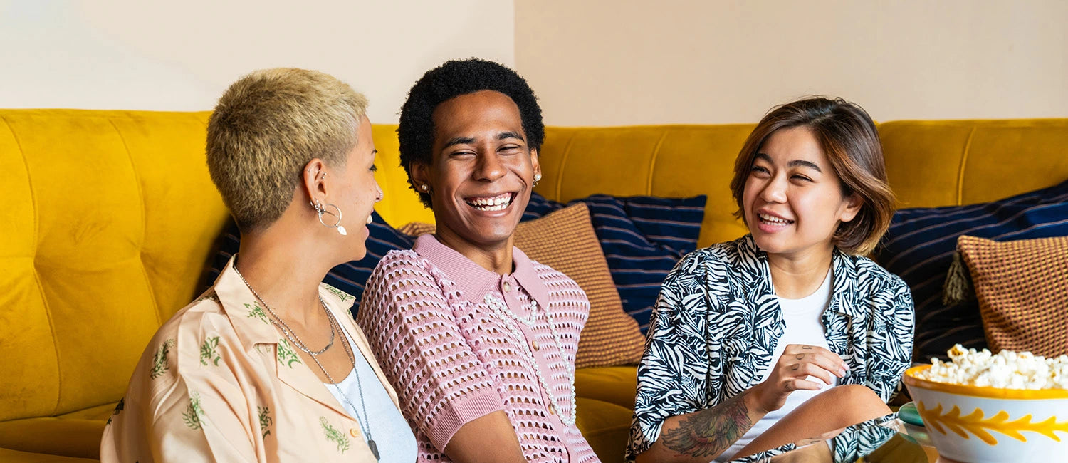Three people sitting on a yellow couch laughing together, with a bowl of popcorn on a table in front of them.